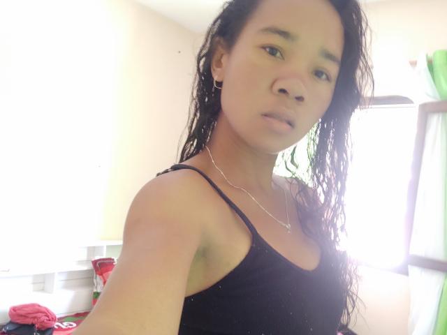 Aniricha hat nasses, schwarzes Haar, ein ernstes Gesicht, kleine Brüste und ein schwarzes Tanktop. Sie trägt eine silberne Halskette mit einem kleinen Kreuz und steht in einem hell erleuchteten Raum..