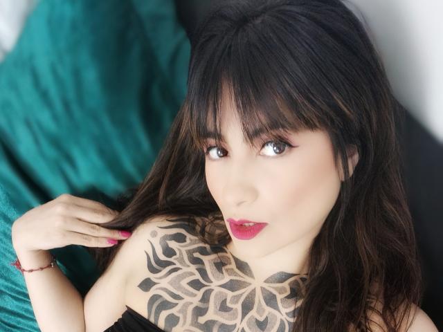 BelleLoren, avec ses cheveux noirs et ondulés et sa frange, son rouge à lèvres rose et son tatouage noir recouvrant son épaule droite, tient ses cheveux avec des ongles roses. Elle regarde l'appareil photo..