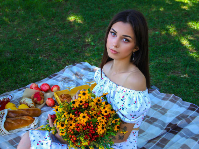 Lara Moss mit langen, braunen Haaren, in einem blumigen Kleid mit Schulterfrei-Ausschnitt, hält einen Sonnenblumenstrauß, rote Beeren und Grünzeug, sitzt auf einer karierten Decke mit Brot, Äpfeln und einem gelben Korb..