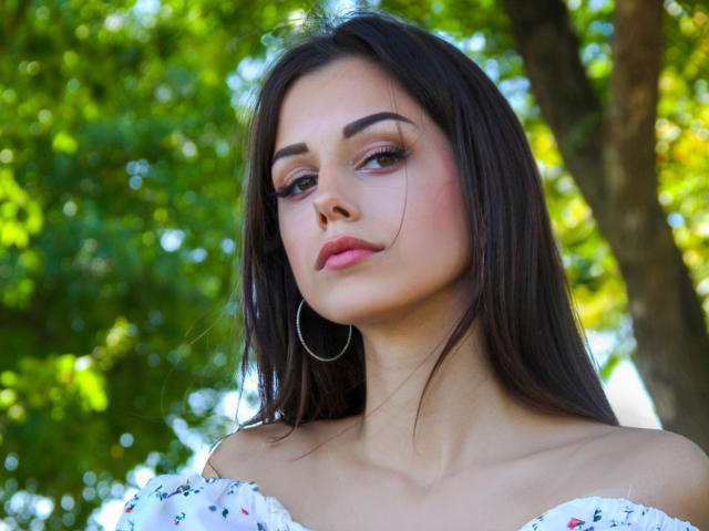Lara Moss a les cheveux brun foncé, des boucles d'oreilles pendantes et un haut blanc à une épaule avec un motif floral. Elle regarde légèrement sur le côté avec une expression subtile et confiante..