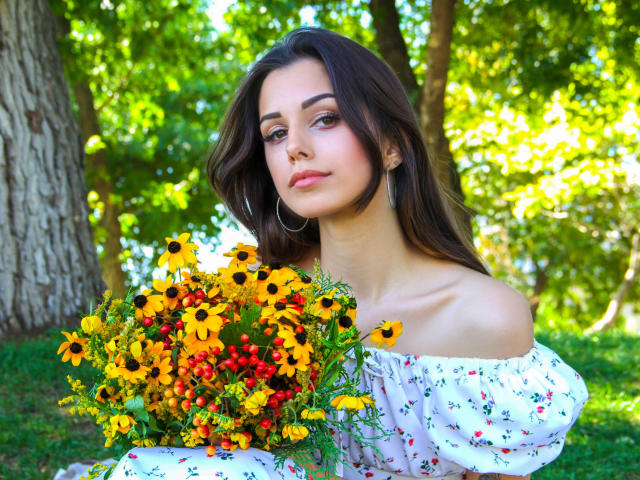 Lara Moss, con lunghi capelli castani, indossa un abito bianco con fiori e spalline scoperte, tiene un bouquet di girasoli, bacche rosse, foglie verdi e sullo sfondo si vede il tronco di un albero. Indossa orecchini a cerchio e guarda leggermente di lato..