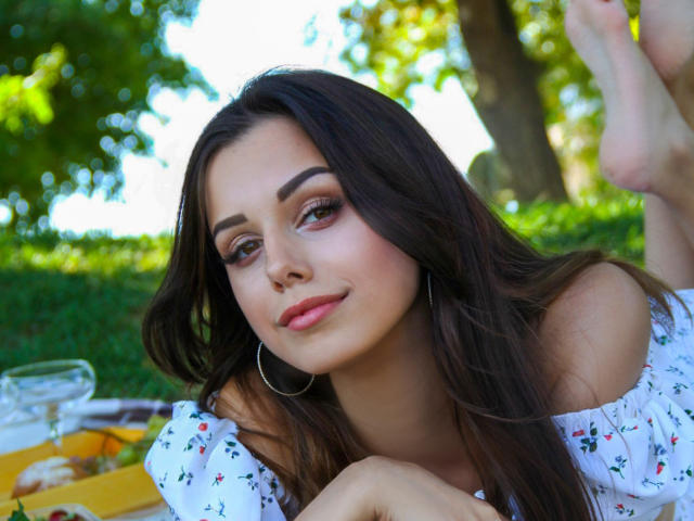 Lara Moss mit langen, braunen Haaren, Reifenohrringen und pinkem Lippenstift, liegt auf dem Gras, trägt ein weißes Kleid mit Blumenmuster und blickt mit einem leichten Lächeln in die Kamera, die Beine angezogen..