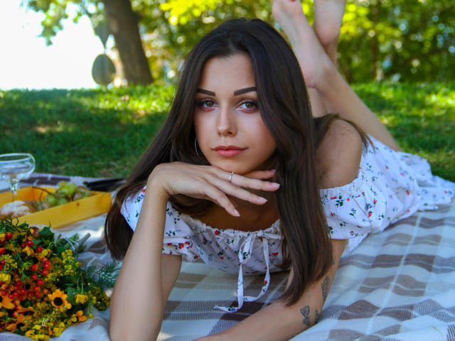 Lara Moss, con el pelo largo y castaño, acostada sobre una manta a cuadros, vestida con un vestido floral, con el mentón apoyado en la mano, las piernas levantadas, mostrando su trasero desnudo, al aire libre..
