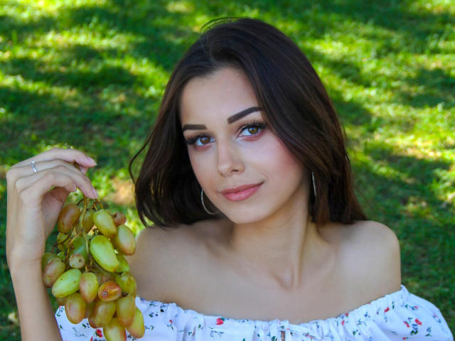 LaraMoss with dark brown hair, holding a bunch of green grapes, wearing a white off-shoulder top, small hoop earrings, and a subtle smile. Her tits are slightly visible.