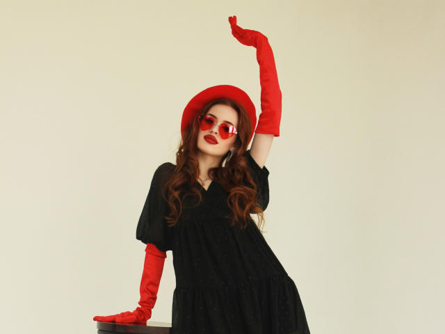 CherylFoxy with red hat, red sunglasses, red gloves, and black dress, posing with arm raised, showing off her tits and red lips, standing against a plain background.