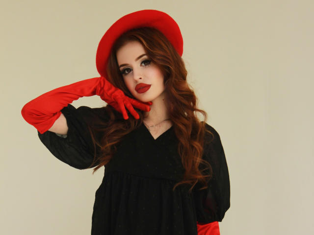 CherylFoxy with wavy red hair, red hat, red gloves, black dress, touching face with gloved hand, red lipstick, pale skin, looking at camera, soft smile, confident.