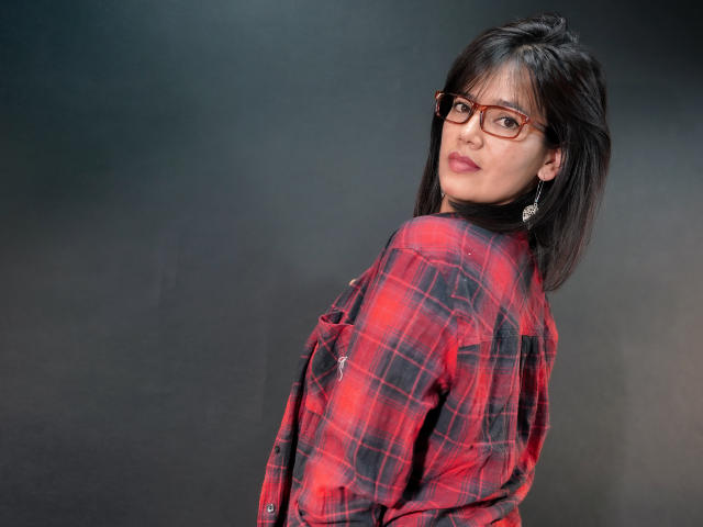 Sashasweetx with brown glasses, red plaid shirt, and earrings, looking back over shoulder, showing her ass, dark hair, and a slight smile.