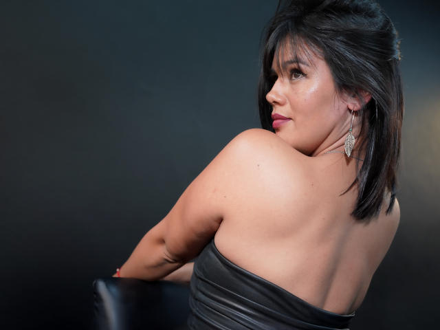 Sashasweetx mit schwarzen Haaren, die über ihre Schulter schaut, trägt ein trägerloses schwarzes Lederkleid, das ihren nackten Rücken und Hintern zeigt, mit Ohrringen und einem leichten Lächeln..