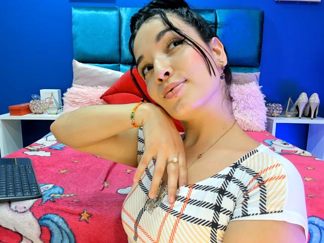 SarahGood with puffy lips, dark hair in a ponytail, plaid shirt, ring on finger, bracelet, lying on pink bed, holding phone, blue wall, red pillow, relaxed, smiling.
