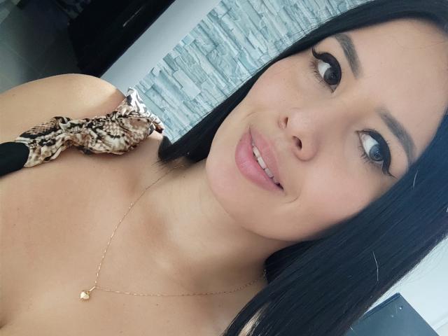 AshleyKatelyn with black hair, brown eyes, and pink lips, wearing a snake-patterned top, necklace with small gold pendant, smiling slightly, looking to the side.