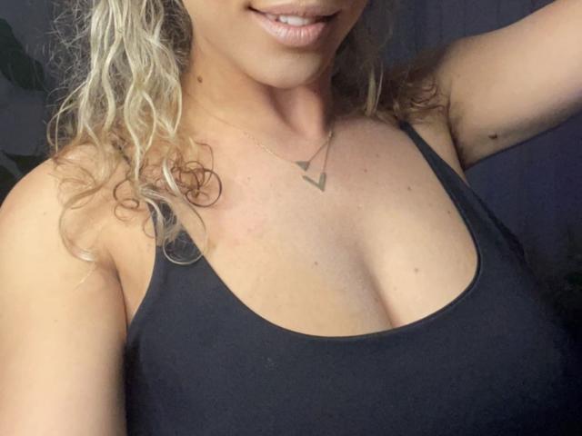 LucilleXSugar met krullend blond haar, lachend, gekleed in een zwart tanktopje, waarbij haar borsten zichtbaar zijn, en met een driehoekige ketting, in een selfie met weinig licht en een lichte onscherpte..