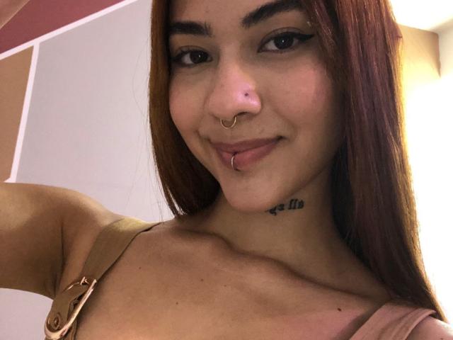 CieloSmith a de longs cheveux bruns, un anneau au nez et un anneau aux lèvres, avec un tatouage sous le menton, et elle tire la langue. Ses seins sont petits. Elle sourit..