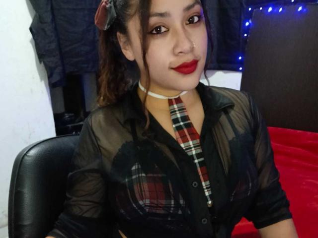 AmelyKent with dark brown hair in a ponytail, wearing a sheer black shirt, plaid tie, red lipstick, and a white choker, sitting on a black chair.