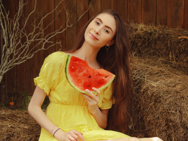 SaraQuincy with long brown hair, yellow dress, holding a slice of watermelon, smiling, wooden fence background, wearing gold hoop earrings, sitting on hay, relaxed, cute.