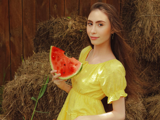 SaraQuincy met lang bruin haar, een glanzende gele jurk, die een plakje watermeloen vasthoudt, een houten omheining, hooibalen, lachend, groene ogen, kleine borsten, lichte huid, oorbellen..