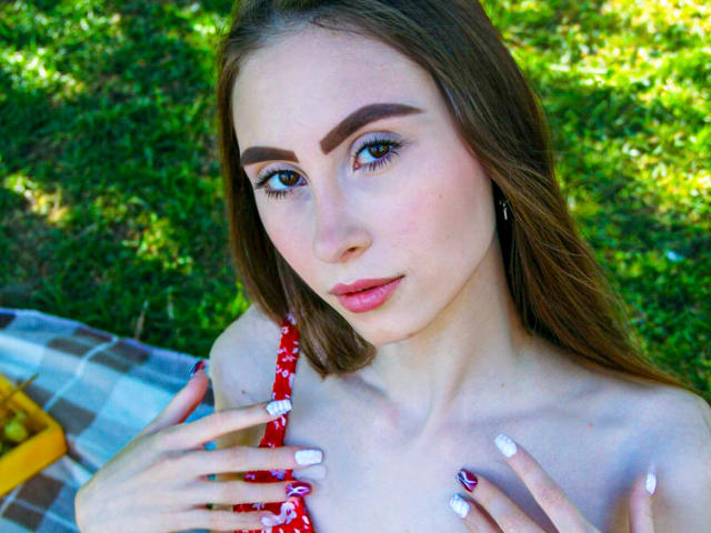 SaraQuincy a la peau claire, les yeux marron et de longs cheveux bruns. Elle est nue sur le torse, avec des ongles blancs, et tient une sangle rouge, montrant ses petites seins. Elle est assise sur une couverture à carreaux..