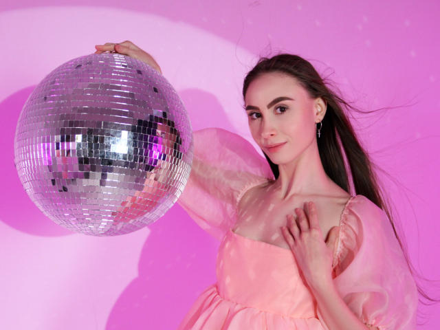 SaraQuincy, avec de longs cheveux bruns, une robe rose, tenant une boule de miroir argentée, fond rose, main droite sur la poitrine, main gauche sur la boule, mignonne, sexy, éclairage rose..