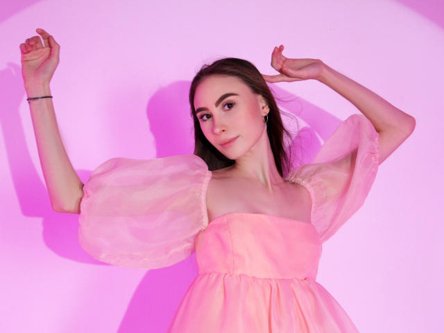 SaraQuincy, vêtue d'une robe rose à manches bouffantes, les bras levés, souriante, peau pâle, cheveux bruns, maquillage discret, petite poitrine, silhouette mince, éclairage rose, bracelet noir..
