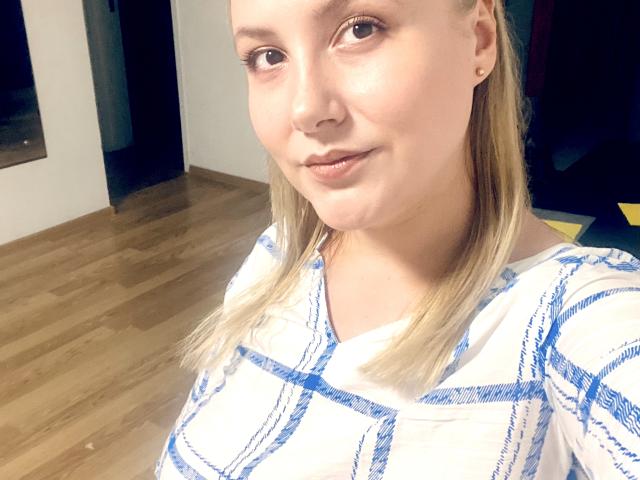 HanterHellen con i capelli biondi, una camicia a quadri bianchi e blu, un sorriso discreto, pelle chiara, piccoli orecchini e uno sfondo con pavimento in legno in un selfie..
