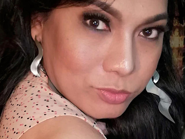 Chanteuse ha i capelli scuri, gli occhi marroni e un leggero sorriso. Indossa una camicia rosa a pois e orecchini argentati a forma di onda. La sua pelle è abbronzata e sta guardando la fotocamera..