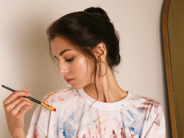 LaraMoss with dark hair in a messy bun, painting her cheek with a brush, wearing a white paint-splattered shirt, looking down, holding a yellow paint in her right hand.