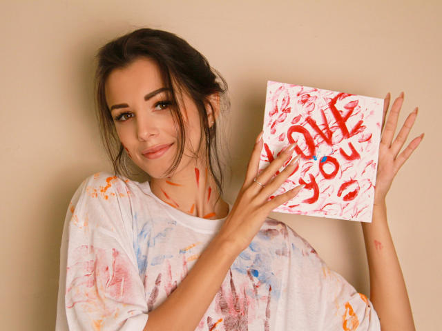 Lara Moss com tinta espalhada nos seios e no peito, segurando um pedaço de papel salpicado de vermelho com a frase "EU TE AMO", vestindo uma camisa branca manchada de tinta, sorrindo..