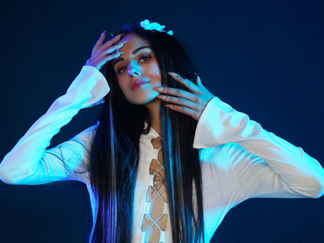 Lara Moss met lang zwart haar, die een witte top met veters draagt, met haar handen voor haar gezicht, blauw en paars licht op haar huid, en een witte bloem in haar haar..