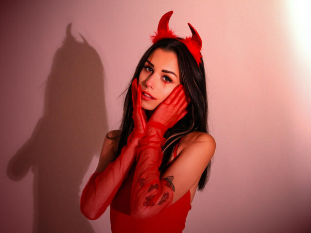 LaraMoss with red devil horns, red gloves, and red dress, hands on face, long black hair, tattoo on right arm, shadow on wall, red lighting, sexy look.