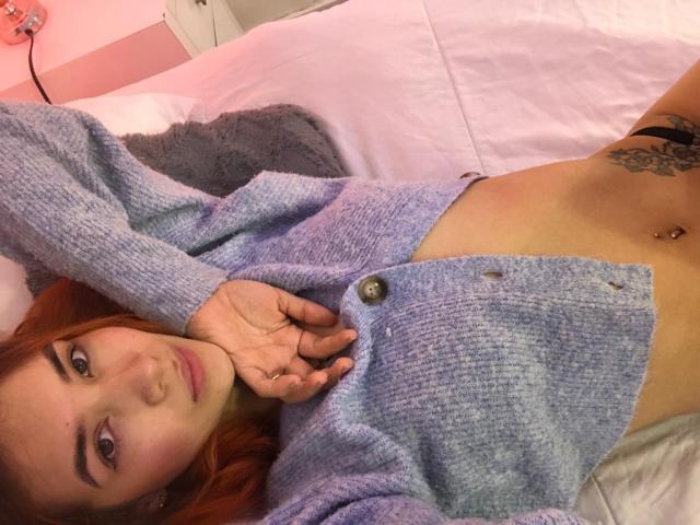 Greta Harris, met rood haar, liggend op bed, gekleed in een paarse cardigan, waarbij haar navel en een tatoeage onder haar navel zichtbaar zijn, met een hand in de buurt van haar gezicht..