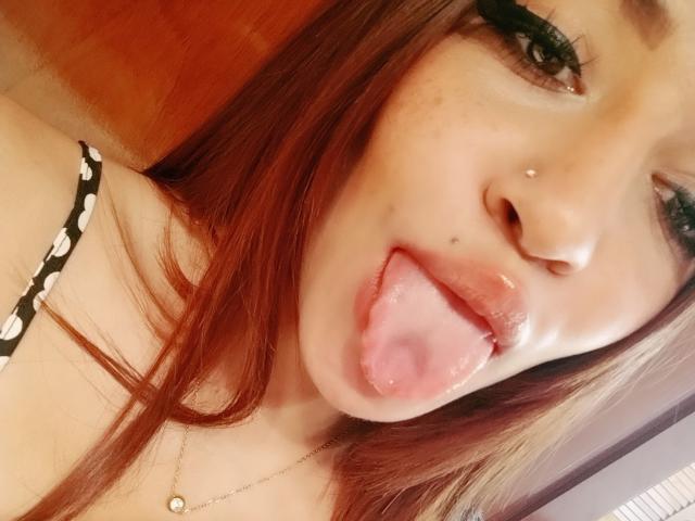 AnnaKhalo, con cabello rojo, piercings en la nariz y la lengua afuera, mostrando su lengua rosa y labios ligeramente entreabiertos. Tiene pestañas largas y un collar de oro..