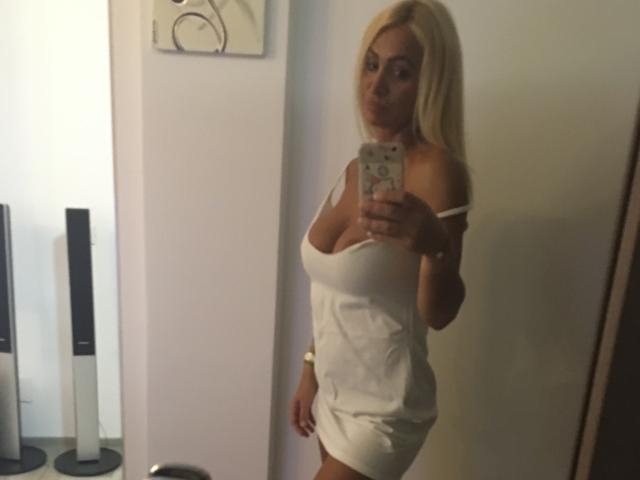Blond BriliantOne with large tits in a tight white dress, taking a mirror selfie, showing her ass, in a dimly lit room with a camera in her hand.