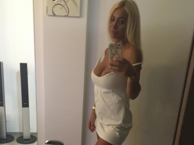 BriliantOne with blonde hair, large tits, wearing a tight white dress, taking a mirror selfie, holding a phone with a floral case, in a dimly lit room.