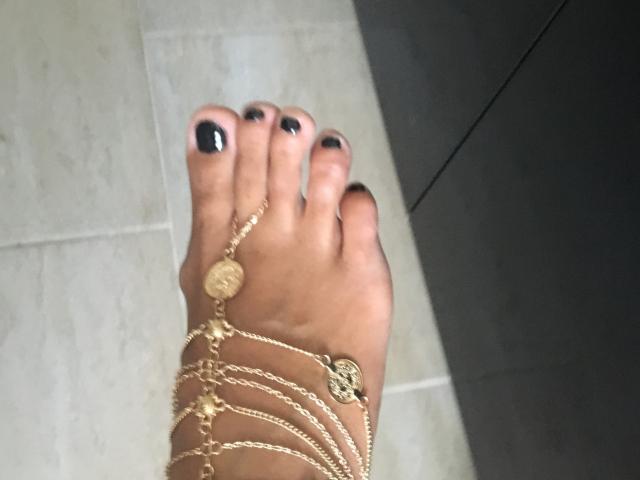 BriliantOne's foot with black-painted toes, adorned with a gold ankle chain featuring small ornaments and a circular charm, standing on a tiled floor.