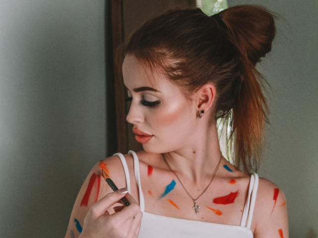 CherylFoxy with painted tits, wearing a white tank top, holding a brush, red, blue, and orange paint on her chest, red lipstick, green scrunchie in her brown ponytail.