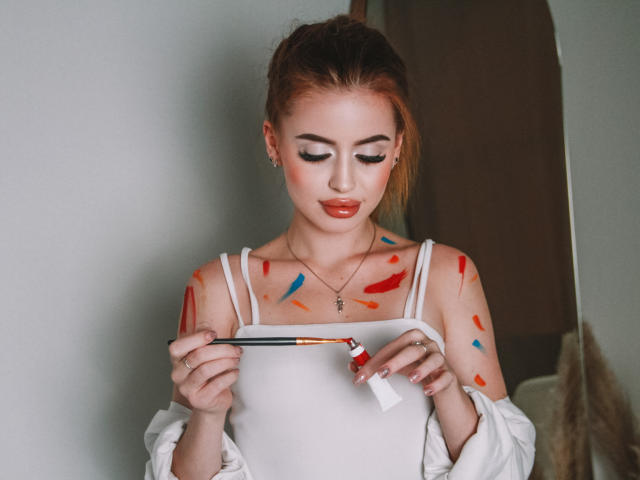 CherylFoxy with painted tits, white top, holding brush, red and blue paint on shoulders, red lipstick, ponytail, necklace, standing, looking down, soft lighting, realistic photograph.