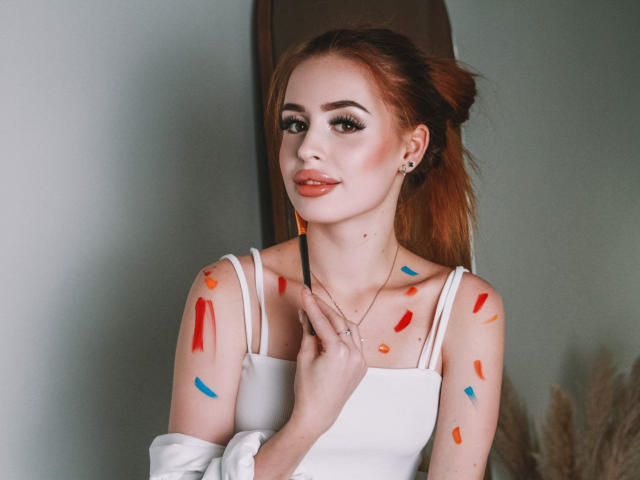 CherylFoxy with painted shoulders, wearing a white top, holding a brush near her neck, has red and blue paint on her skin, and a subtle smile, with red lipstick.