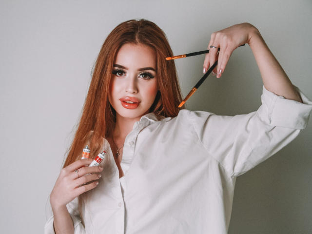 CherylFoxy with long red hair, wearing a white shirt, holding makeup brushes in one hand and a makeup tube in the other, looking at the camera with a slight smile.