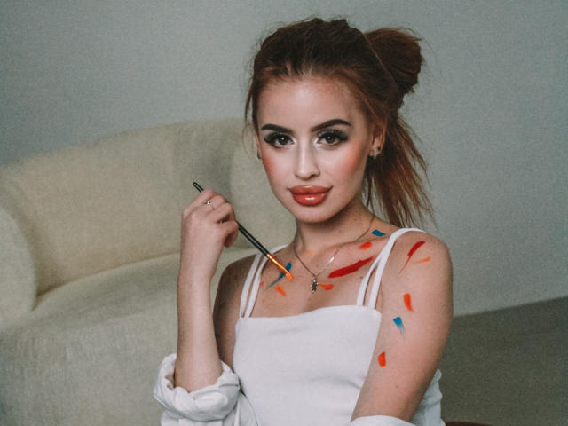 CherylFoxy with painted tits, wearing a white off-shoulder top, holding a paintbrush, has red and blue paint on her chest and arm, looking at the camera.
