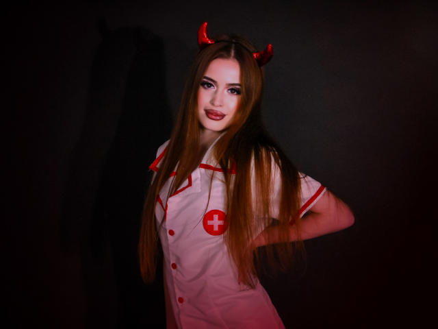 CherylFoxy with long brown hair, red devil horns, white nurse outfit, red cross, hands on hips, dark background, red makeup, smiling, cute, sexy.