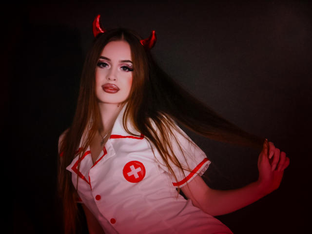 CherylFoxy with long brown hair, red devil horns, white nurse outfit, red cross, holding hair, dark background, red lighting, looking at camera, smiling, full lips, pale skin.