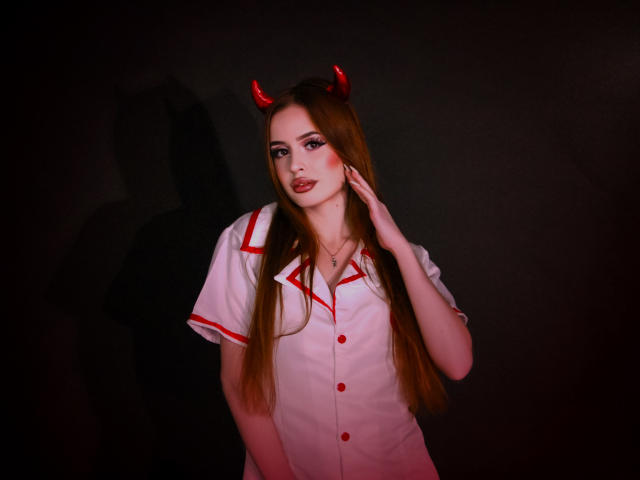 CherylFoxy with long brown hair, wearing a white nurse outfit with red trim, red devil horns, and red buttons, touching her face, looking seductively at the camera.