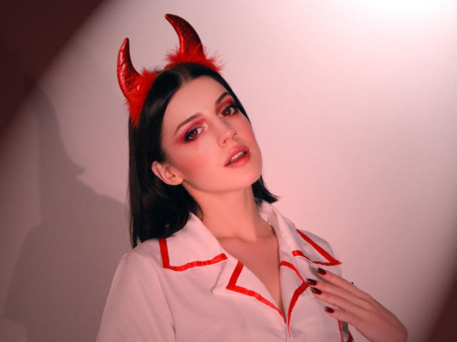 MiaFane, avec des cornes de diable rouges, portant une chemise blanche avec des détails rouges, a les cheveux noirs, du fard à paupières rouge et un anneau de nez, sa main sur sa poitrine, regardant la caméra..