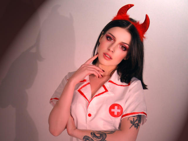 MiaFane avec des cornes de diable rouges, une tenue de nurse blanche avec des détails rouges, une croix rouge, des cheveux noirs, des tatouages sur les bras, un doigt sur la joue, une ombre sur le mur, regardant la caméra..