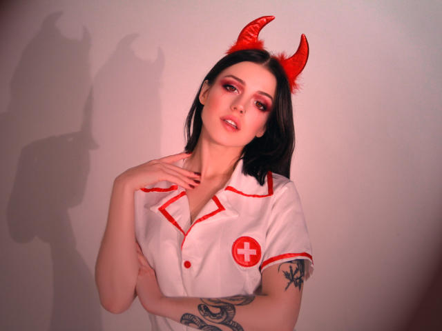 MiaFane avec des cornes de diable rouges, une tenue de nurse blanche, une croix rouge, du maquillage rouge, des cheveux noirs, un tatouage sur le bras droit, une main sur la poitrine, une ombre sur le mur. Seins visibles, sexy..