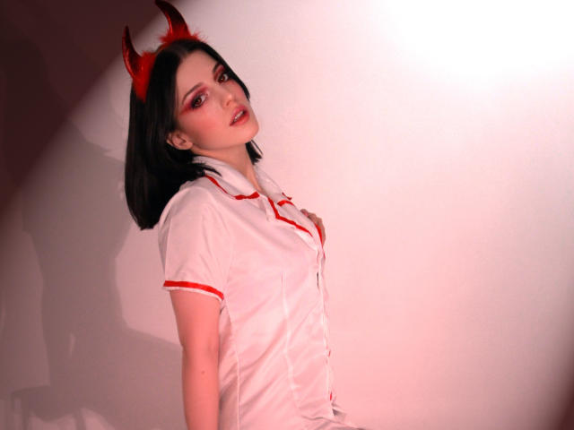 MiaFane avec des cornes de diable rouges, une chemise blanche à boutons avec des détails rouges, une coupe de cheveux courte noire, et un fard à paupières rose, debout de côté, regardant la caméra avec un léger sourire..
