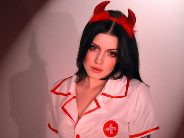 MiaFane avec des cornes de diable rouges, des cheveux noirs, une tenue de nurse blanche avec des détails rouges, une croix rouge, des yeux rouges, regardant la caméra, une ombre sur le mur, sexy, mignonne, lumière douce..