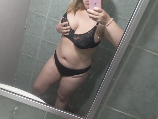 HotNata, con i capelli biondi, indossa un reggiseno di pizzo nero e mutandine nere, mostrando i suoi seni e il sedere, mentre scatta un selfie allo specchio in un bagno con piastrelle verdi, tenendo in mano un telefono rosa..