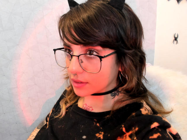 AmySpider met kattenoren, een bril, een neusring, oorringen, een zwarte choker, tatoeages en golvend bruin haar, gekleed in een zwart shirt met een vlammenpatroon, kijkt naar de zijkant..