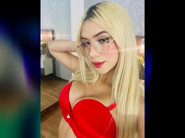 ChristieGarci with blonde hair, glasses, and red bra, showing off her big tits. She's got a cute face, red lipstick, and a confident smile.