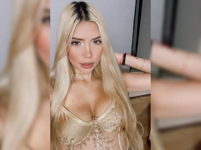 ChristieGarci with long blonde hair, wearing a sheer, gold lace bralette, showing her big boobs, looking in a mirror with her hand behind her head.